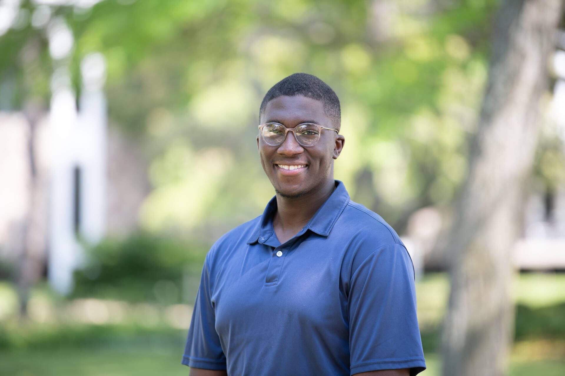An individual smiling at the camera with a trees and foliage in the background.